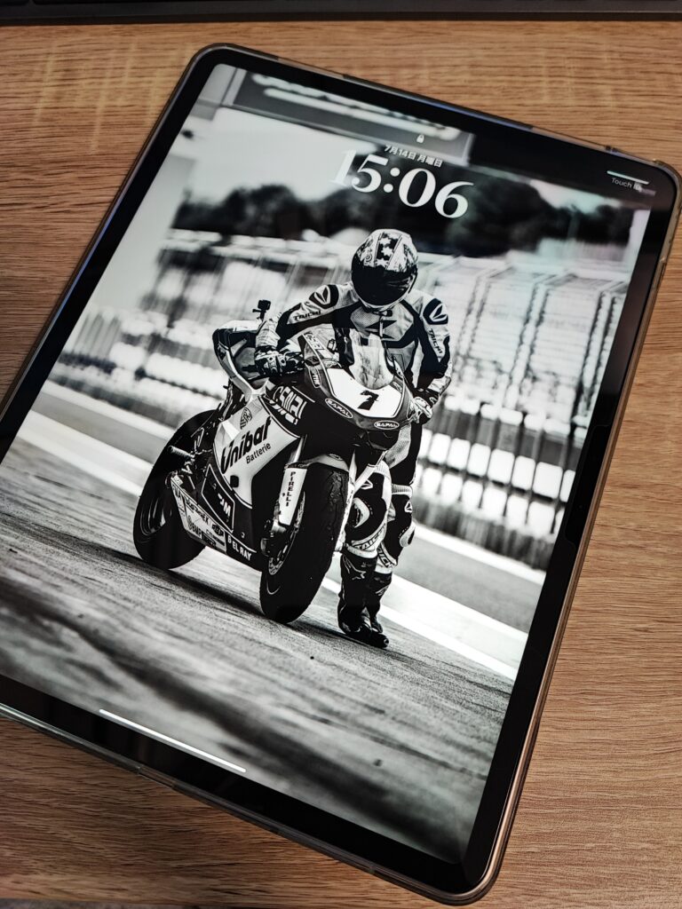 木目のテーブル上に置かれたiPad。ロック画面にはモノクロ調のバイクレーサーの写真が表示されている。
