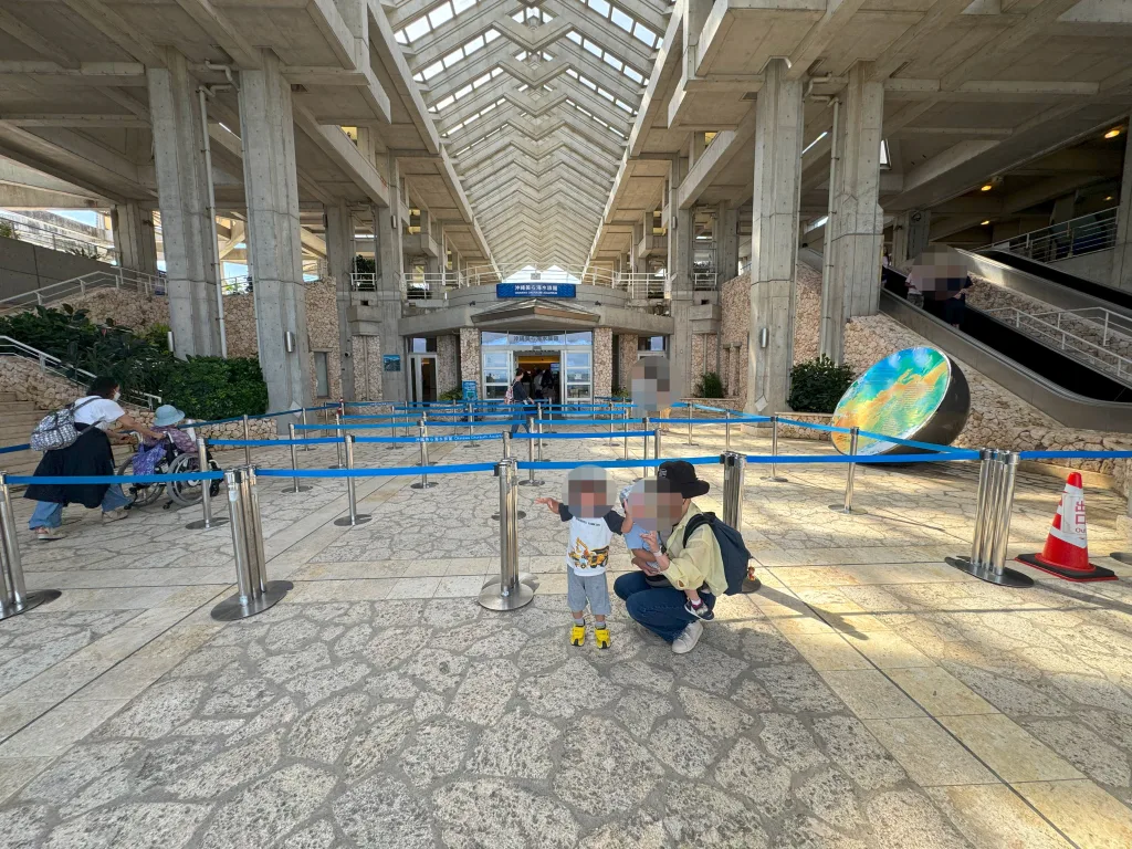 沖縄美ら海水族館のエントランス前で記念撮影する家族。コンクリート造りの建物と青い案内サインが特徴的。