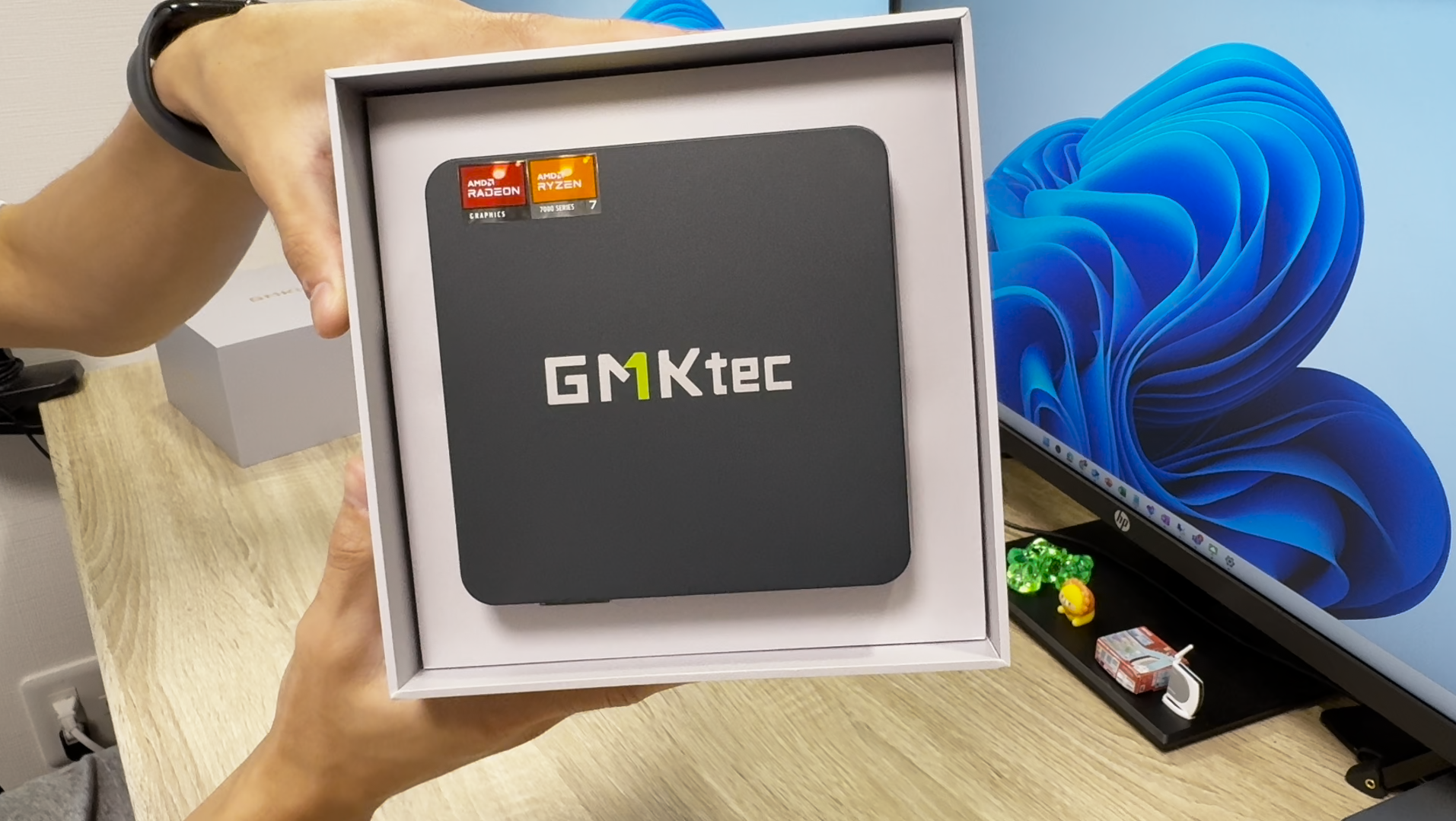 GMKtecのミニPC「NucBox K6」が箱に収められた状態で手に持たれている様子。背景にはWindows 11のデスクトップ画面