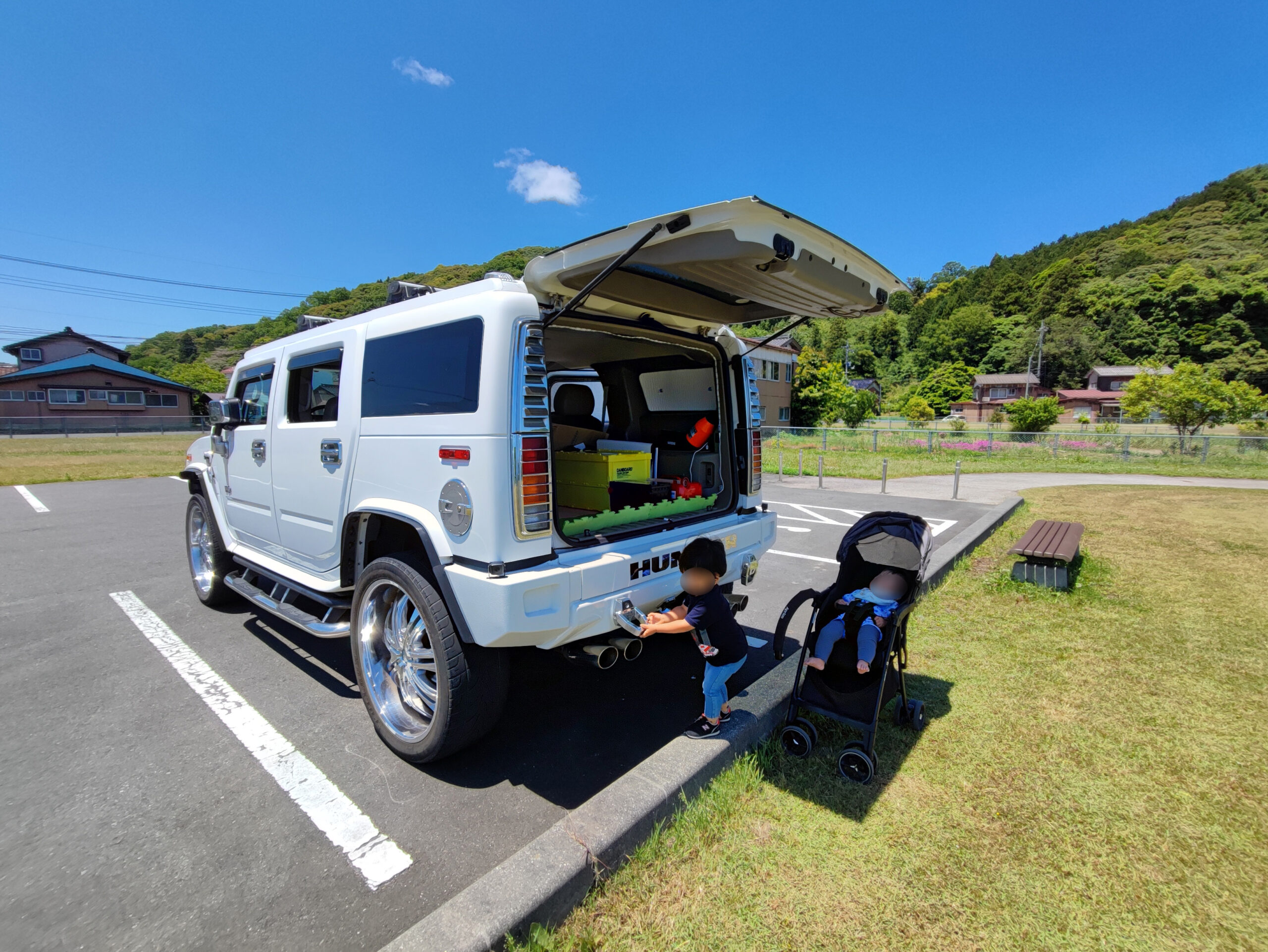 白いハマーH2の後部で遊ぶ子どもとベビーカー、青空の下の駐車場にて撮影