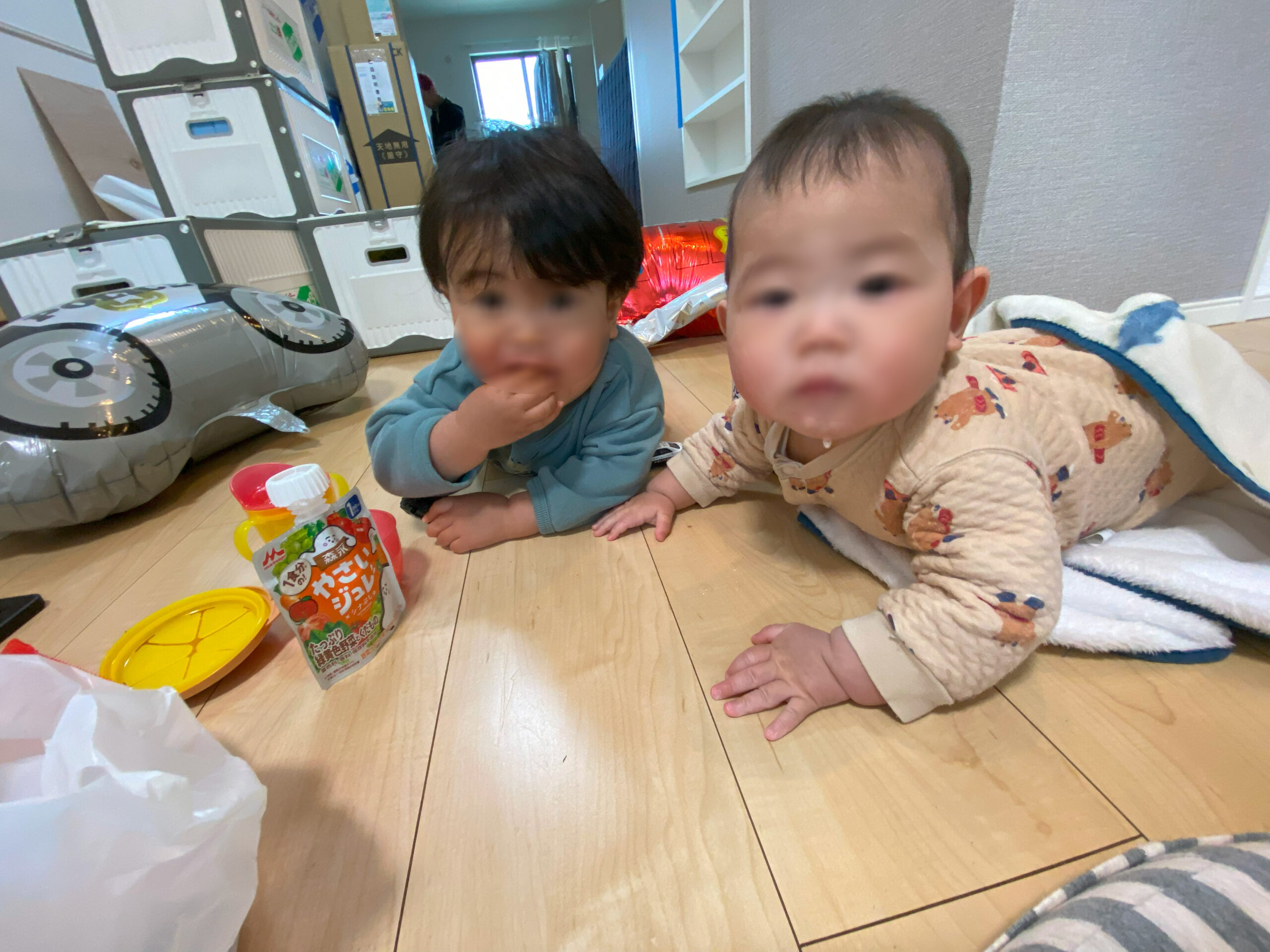 引っ越し作業中の室内で、床に座って遊ぶ二人の幼児。背景には荷物や段ボールが積まれている様子が見える。