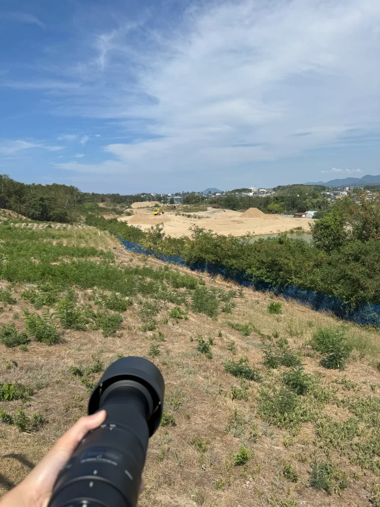 望遠レンズで建設現場を遠望している様子。広がる青空と工事中の砂地が印象的。