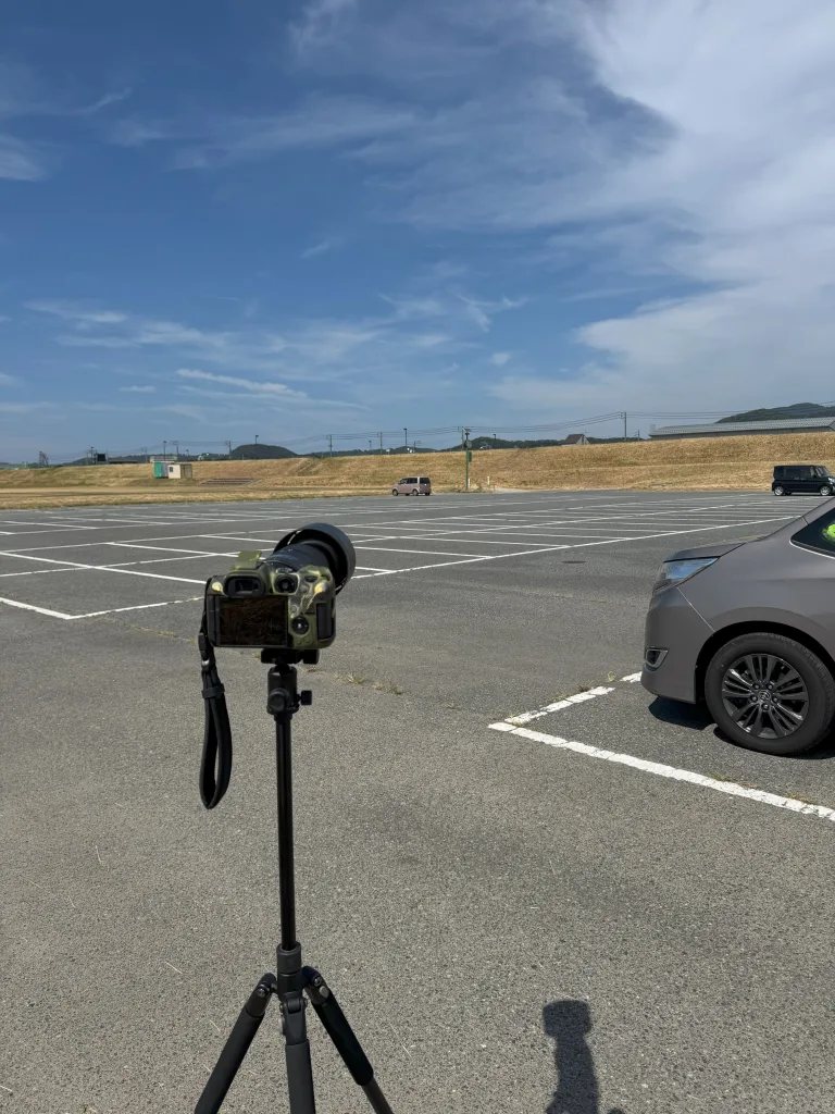 青空の下、空いた駐車場に設置された三脚とカモフラージュ柄のカメラ。背景には車と遠くの丘が見える。