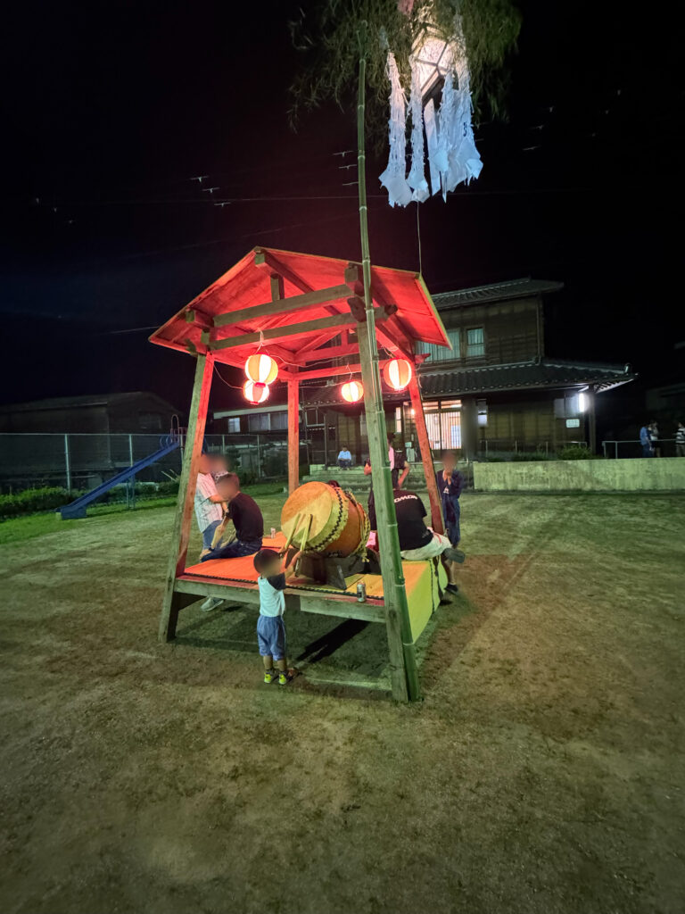 夜の公園で行われている地元の盆踊り。櫓の上で太鼓を叩く人と、提灯の明かりに照らされた子どもたちの様子。
