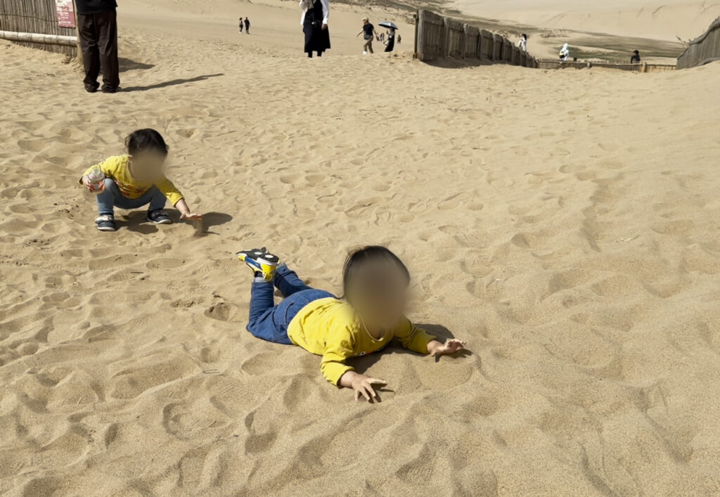 鳥取砂丘で砂の上に寝転びながら遊ぶ子どもたち。青空の下で砂遊びを楽しむ様子。